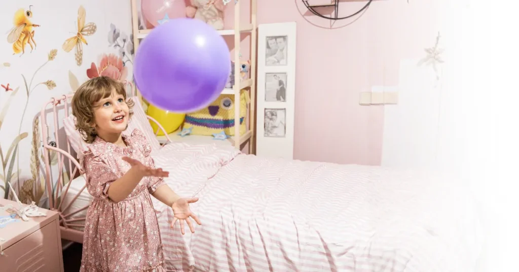 A young child in a pink dress smiles and tosses a purple balloon in a pastel-coloured bedroom with a bed, framed photos, and butterfly wall art.