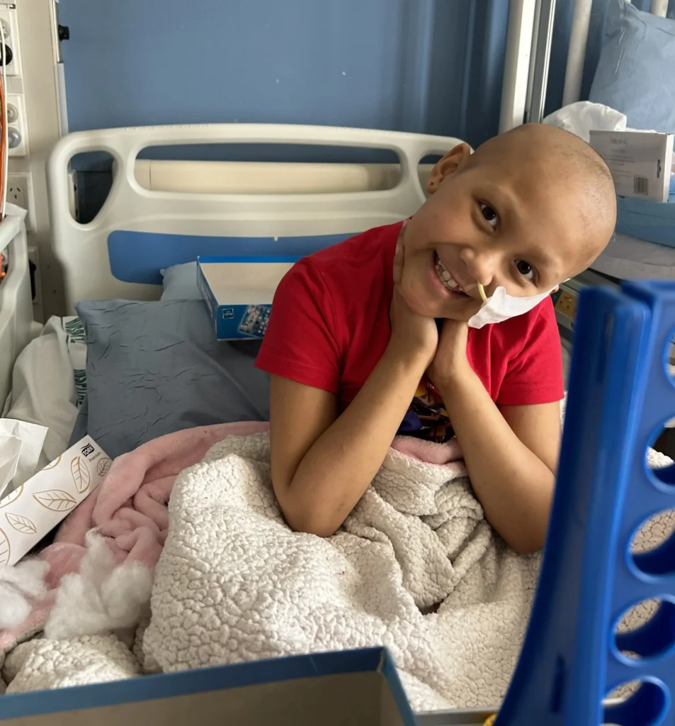A young child with a nasal tube sits up in a hospital bed, smiling and resting their chin on their hands. The child is wrapped in a cosy blanket, wearing a red shirt.