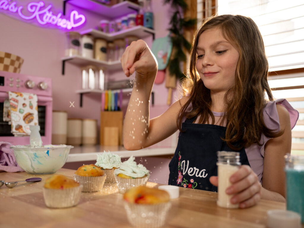 At her new baking station, a girl wearing a personalised apron sprinkles sugar onto freshly iced cupcakes in a kitchen.