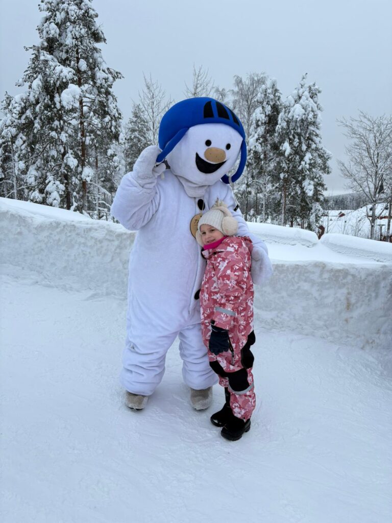 A young child in a pink snowsuit hugs a large snowman mascot wearing a blue helmet in a snowy forest, perhaps imagining a future baking station.