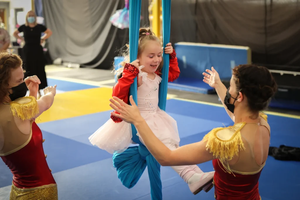 A young girl in a pink dress smiles while swinging on blue aerial silks, assisted by two adults in red costumes and masks, in a gym with mats.