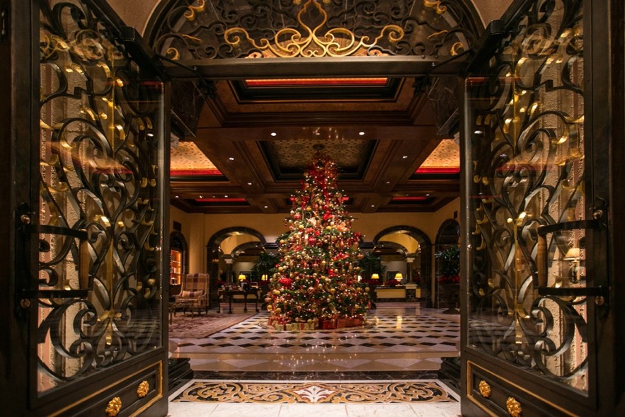 A large, decorated Christmas tree stands in the centre of an elegant hotel lobby, viewed through ornate, open wrought-iron double doors.
