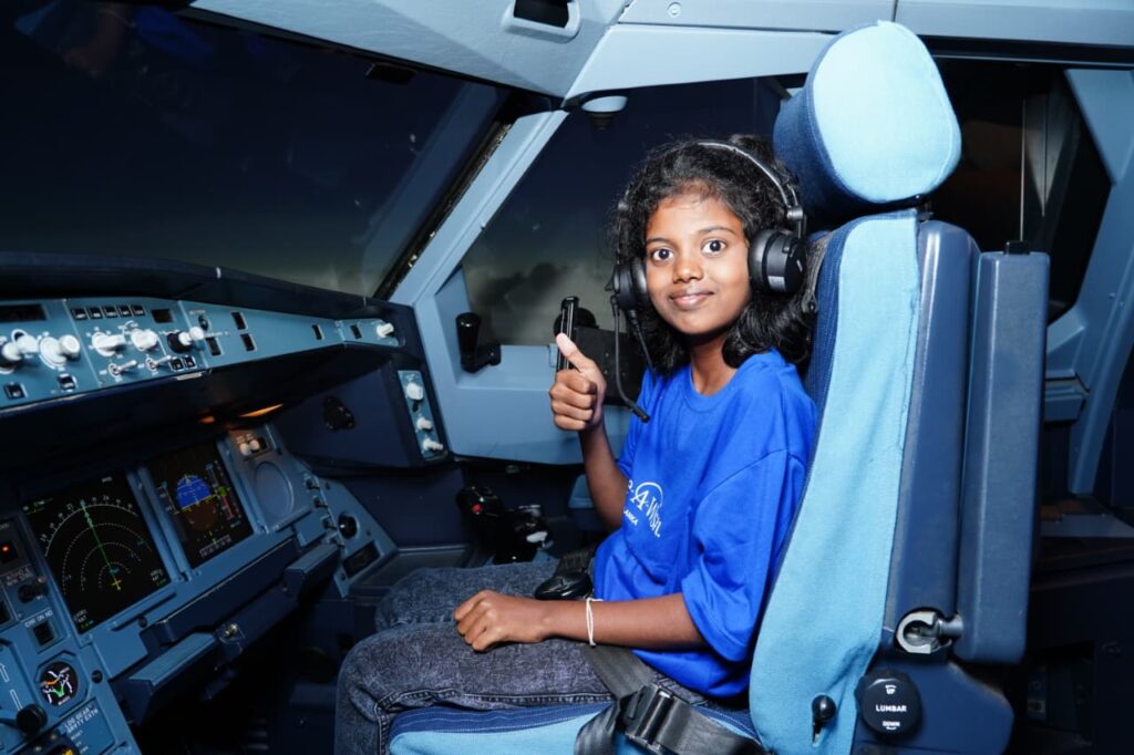 A young girl wearing headphones and holding a soft elephant toy sits in an aeroplane cockpit and gives a thumbs up.