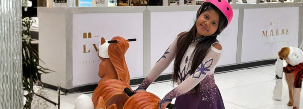 A young girl wearing a pink helmet and purple ice skating dress rides a squirrel-shaped coin-operated kiddie ride in a shopping centre, fulfilling Mía’s wish to go ice skating.
