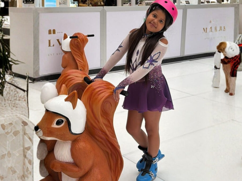 A young girl in a purple skating outfit and pink helmet stands in a shopping centre behind a squirrel-shaped ride, fulfilling Mía’s wish to go ice skating.
