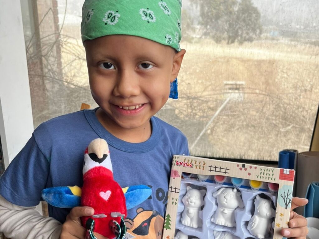 A child wearing a green bandana smiles while holding a soft toy parrot and a craft kit containing three unpainted animal figurines, perfect for Mia’s wish to be a vet.