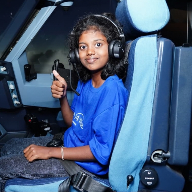 A young girl wearing a blue shirt and headset sits in an aeroplane cockpit seat, smiling and giving a thumbs-up gesture.