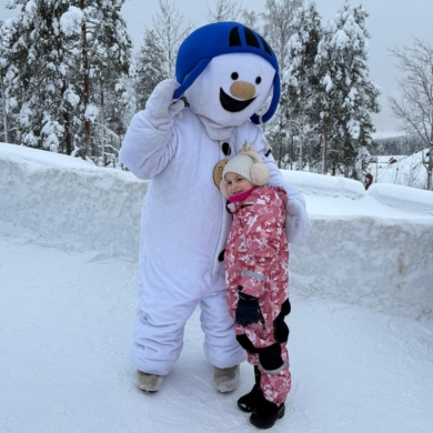 A person in a snowman costume with a blue helmet stands outside in the snow, posing with a child in a pink and white snowsuit. Unwrap the Power of a Wish amid snow-covered trees and a snowy wall behind them.
