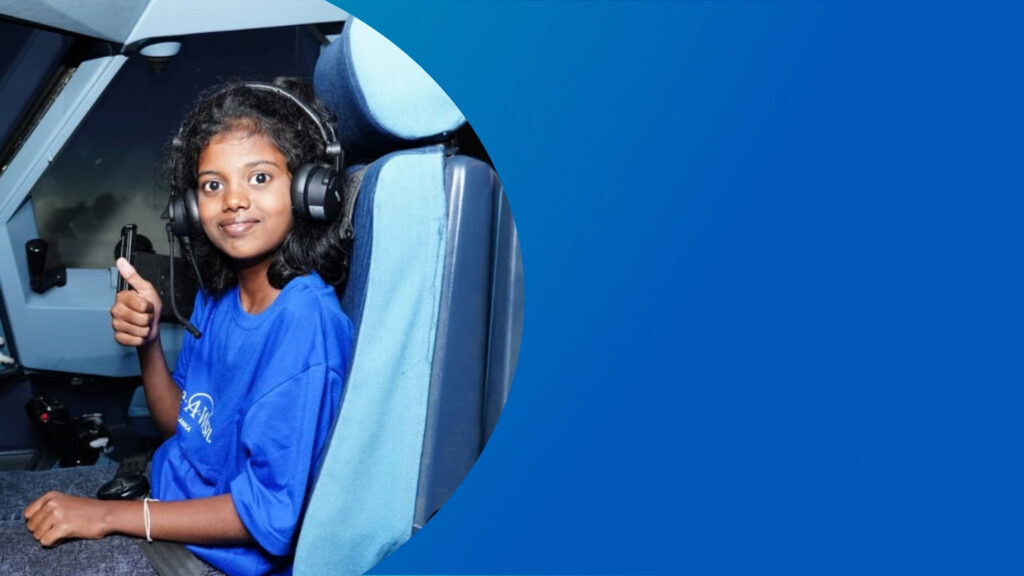 A young girl wearing headphones sits in an aeroplane cockpit seat and gives a thumbs-up.