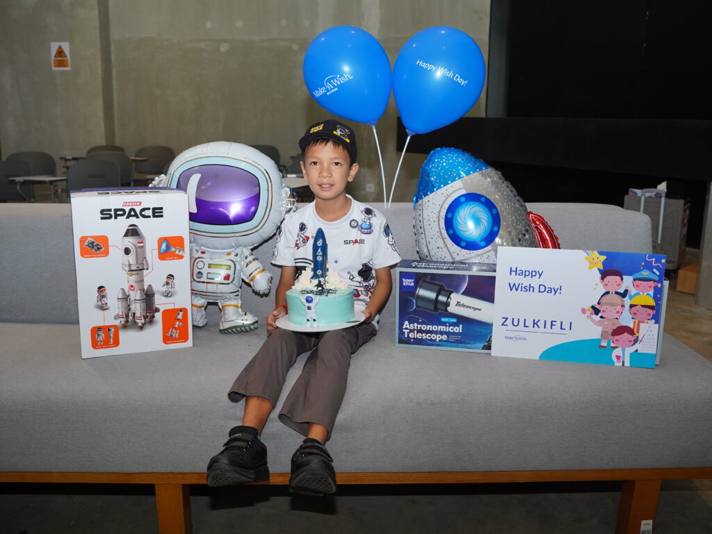 A young boy sits on a grey sofa holding a space-themed birthday cake and a soft elephant toy, surrounded by space-themed presents, a telescope, a sign saying Happy Wish Day! Zulkifli, and two blue balloons.