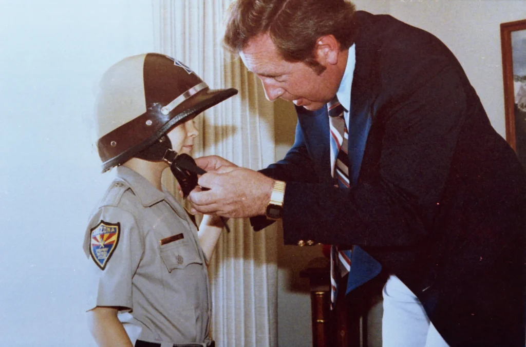 A man in a suit helps a child wearing an Arizona Highway Patrol uniform fasten the chin strap of a motorbike helmet.