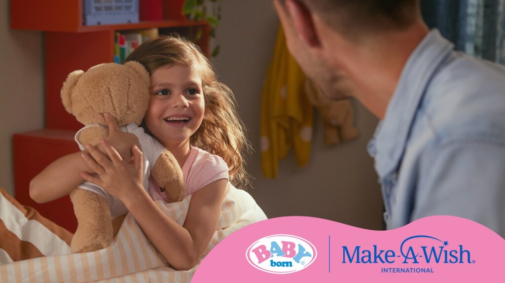 A smiling young girl hugs a teddy bear whilst looking at a man, marking the launch of Make-A-Wish in the Dominican Republic, with Baby Born and Make-A-Wish International logos at the bottom.